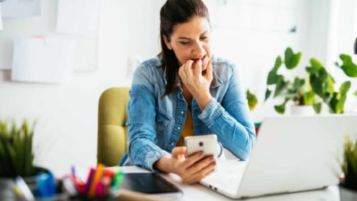 A worried woman looks at her phone and laptop, seeking ways to tighten her belt against inflation.