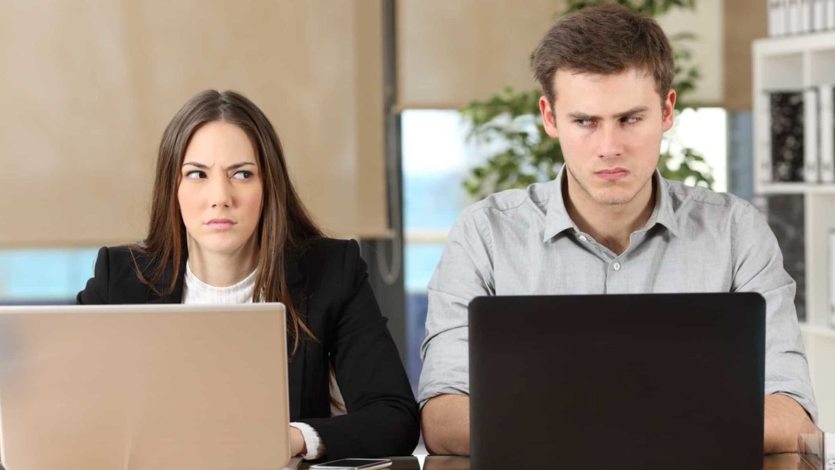 A businesswoman and businessman look sideways at each other during a dispute at their laptops.