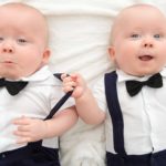 Two twin babies dressed in bow ties, white shirts and braces lie side by side with one grabbing the over shoulder brace of his brother and smiling cheekily at the camera.