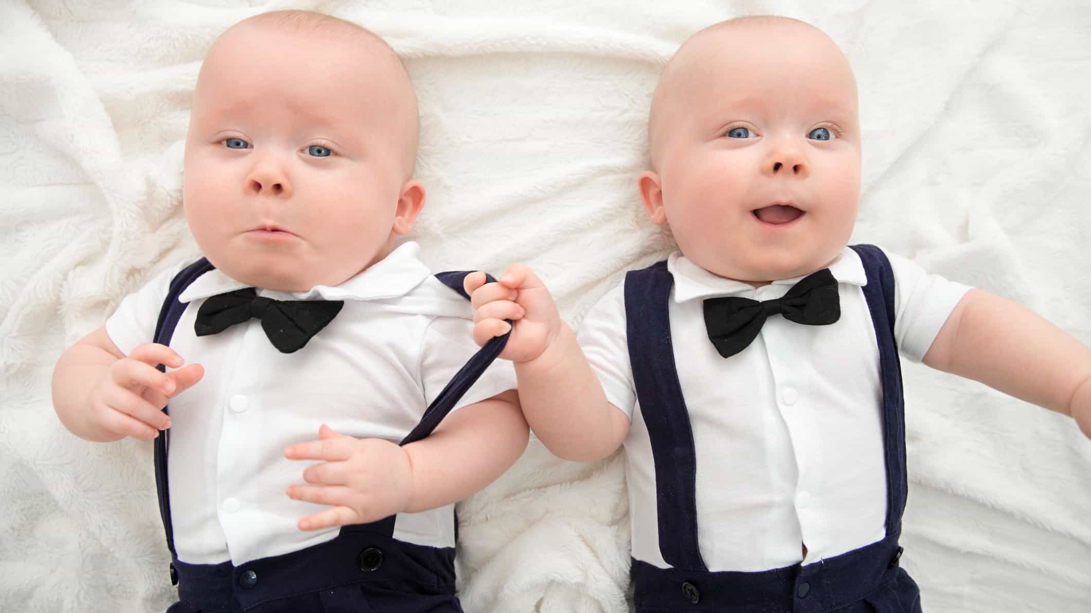 Two twin babies dressed in bow ties, white shirts and braces lie side by side with one grabbing the over shoulder brace of his brother and smiling cheekily at the camera.
