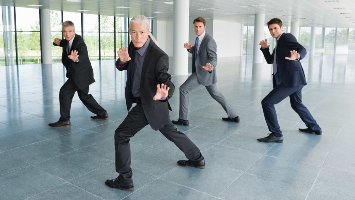 Four businessmen in suits pose together in a martial arts style pose as if ready to engage in competition or spring into a fight.