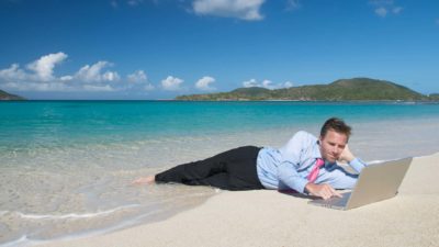 A man in business pants, a shirt and a tie lies in the shallows of a beautiful beach as he consults his laptop on the shore, just out of the water's reach.
