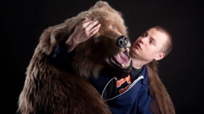 A young man has a look of alarm on his face as he turns to see the close up face of a brown grizzly bear that is draped over him as part of a large life-size bear skin rug he is wearing over his shoulders.