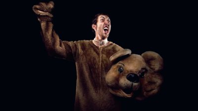A man in a brown bear costume holds the head of it in one hand while raising his other arm in excited victory-style pose.