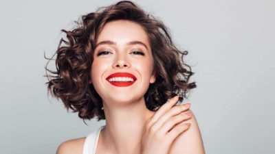 A happy beautiful woman with curly brown hair and wearing bright red lipstick smiles representing the soaring Adore Beauty share price today