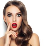 A beautiful woman with brown hair and wearing bright red lipstick looks shocked as she holds her hand to her cheek.