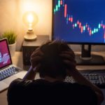 A man sits at his computer with his head in his hands while his laptop screen displays a Bitcoin symbol and his desktop computer screen displays a steeply falling graph.