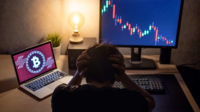 A man sits at his computer with his head in his hands while his laptop screen displays a Bitcoin symbol and his desktop computer screen displays a steeply falling graph.