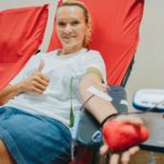 A woman reclines in a comfortable chair while she donates blood holding a pumping toy in one hand and giving the thumbs up in the other as she is attached to a medical machine to collect her blood donation.