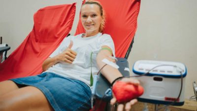 A woman reclines in a comfortable chair while she donates blood holding a pumping toy in one hand and giving the thumbs up in the other as she is attached to a medical machine to collect her blood donation.