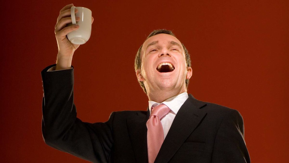 A man in a business suit holds his coffee cup aloft as he throws his head back and laughs heartily.