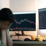 a woman sits next to her computer screen with her head in her hands with the screens slowing graphs on downward trajectories.