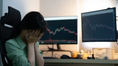 a woman sits next to her computer screen with her head in her hands with the screens slowing graphs on downward trajectories.