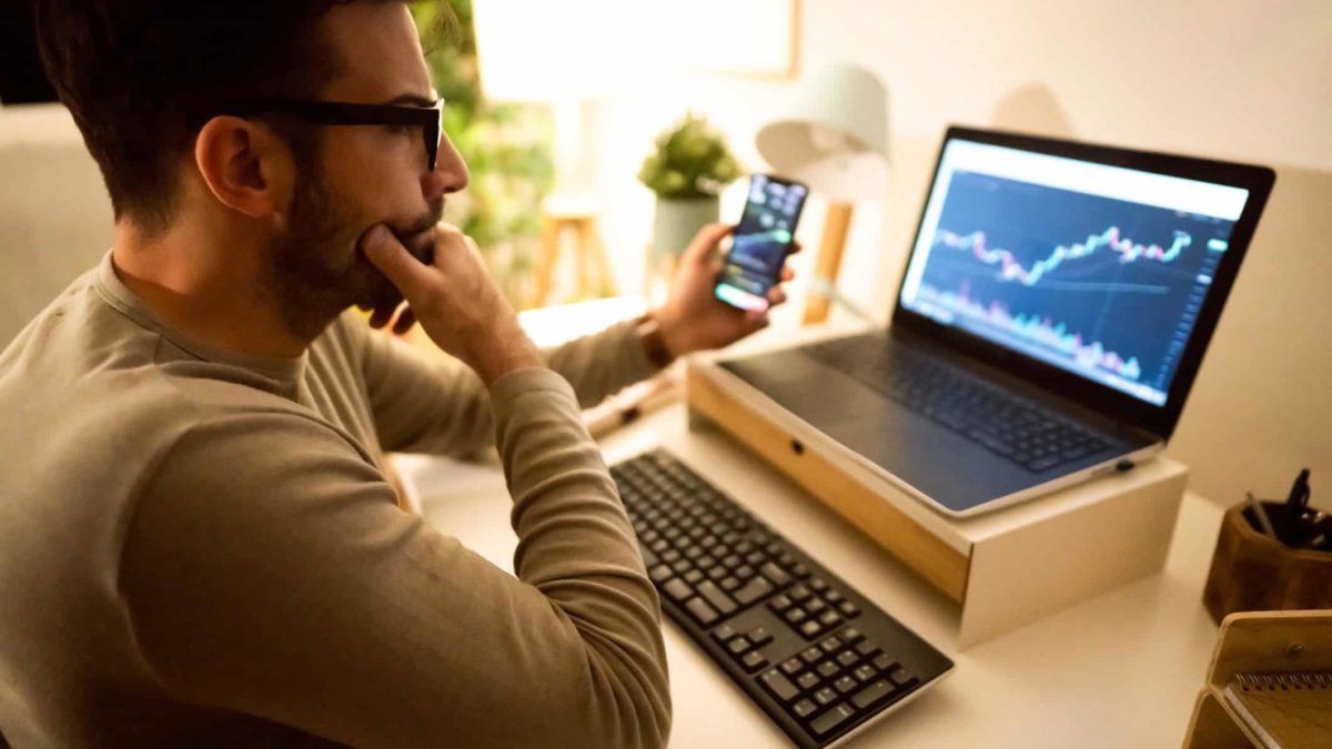 A man sits at a desk with a phone in one hand, his other hand on his chin and studies a computer screen in front of him with what appears to be cryptocurrency data on both screens.