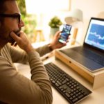 A man sits at a desk with a phone in one hand, his other hand on his chin and studies a computer screen in front of him with what appears to be cryptocurrency data on both screens.