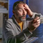 A man sits wide-eyed at a desk with a laptop open and holds one hand to his forehead with an extremely worried look on his face as he reads news of the Bitcoin price falling today on his mobile phone