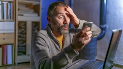 A man sits wide-eyed at a desk with a laptop open and holds one hand to his forehead with an extremely worried look on his face as he reads news of the Bitcoin price falling today on his mobile phone