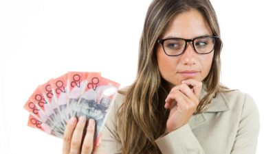 A woman has a thoughtful look on her face as she studies a fan of Australian 20 dollar bills she is holding on one hand while he rest her other hand on her chin in thought.