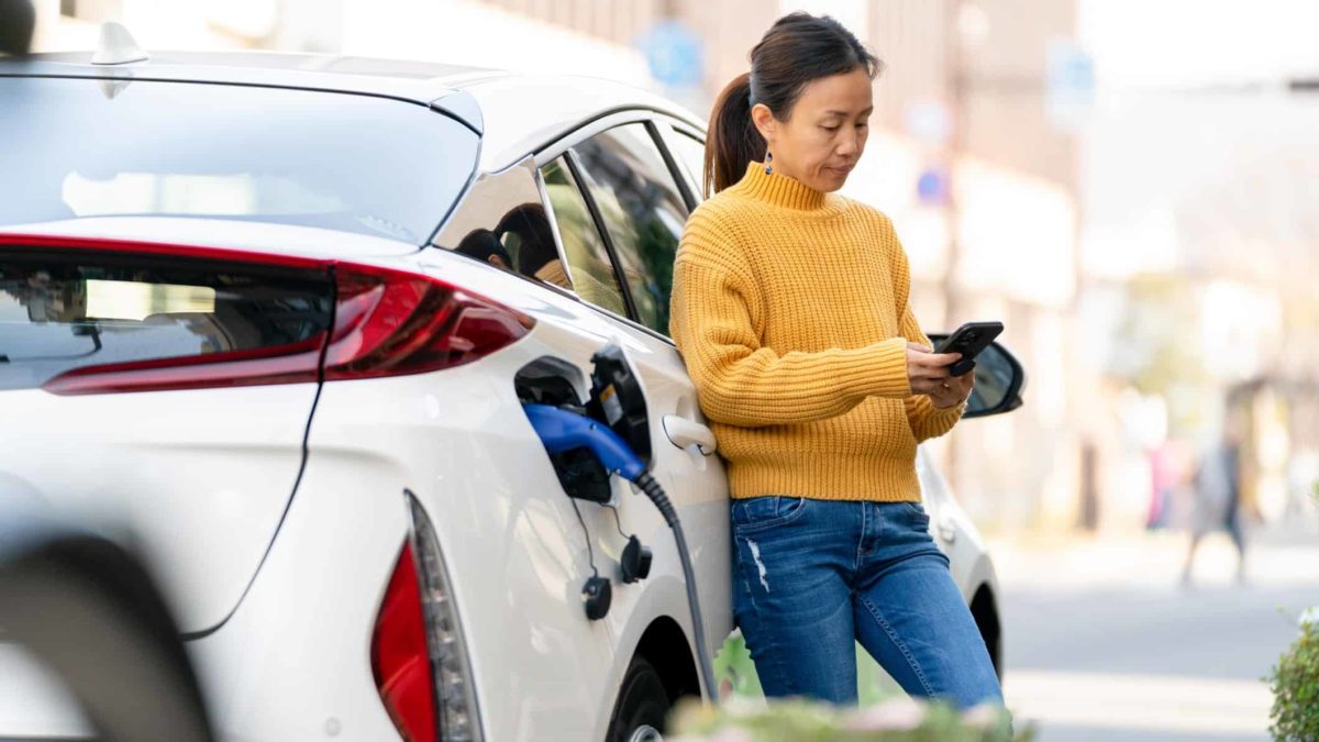 A woman in jeans and a casual jumper leans on her car and looks seriously at her mobile phone while her vehicle is charged at an electic vehicle recharging station.