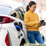 A woman in jeans and a casual jumper leans on her car and looks seriously at her mobile phone while her vehicle is charged at an electic vehicle recharging station.