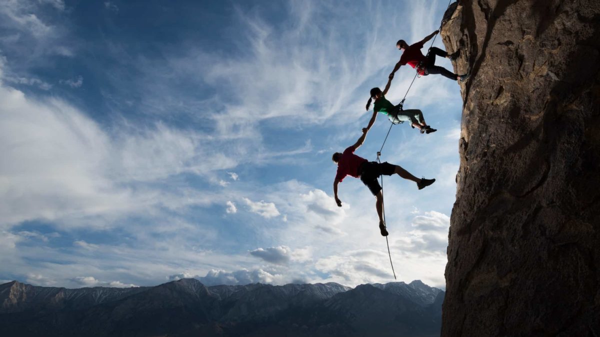 Three rock climbers hang precariously off a steep cliff face, each connected to the other with the higher person holding on and the two below them connected by their arms and rope but not making contact with the cliff face.