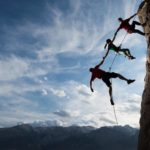 Three rock climbers hang precariously off a steep cliff face, each connected to the other with the higher person holding on and the two below them connected by their arms and rope but not making contact with the cliff face.