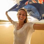 A fresh-faced young woman holds an Australian flag aloft above her head as she smiles widely.
