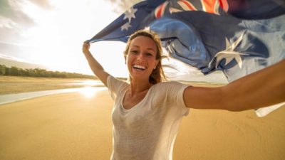 A fresh-faced young woman holds an Australian flag aloft above her head as she smiles widely on a beach as though celebrating a national day or event where Australia has been successful.