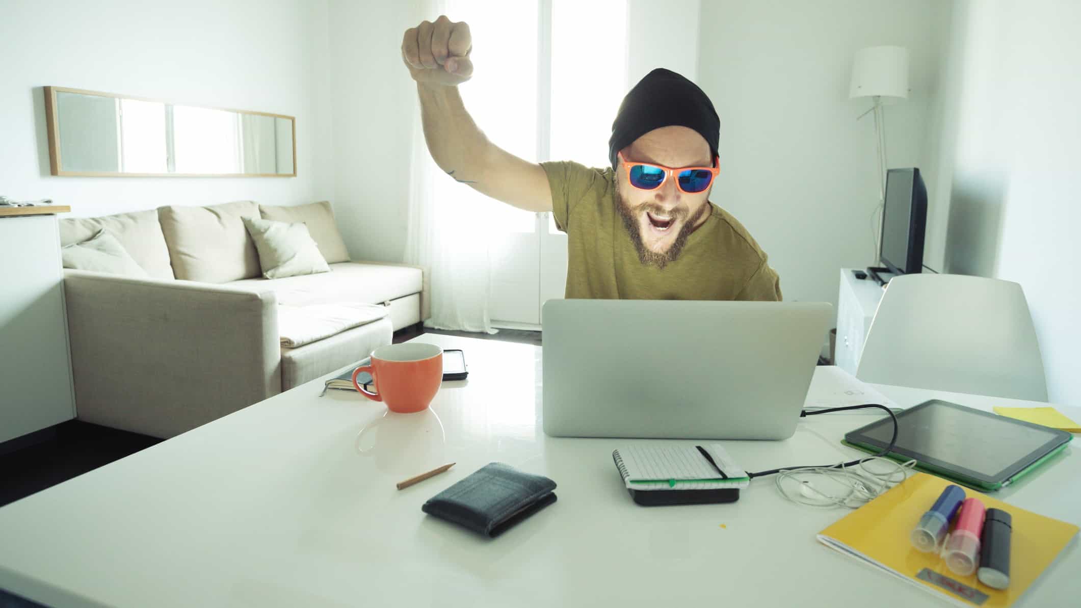 A man with a scrappy beard and wearing dark sunglasses and a beanie head covering raises a fist in happy celebration as he sits at is computer in a home environment.