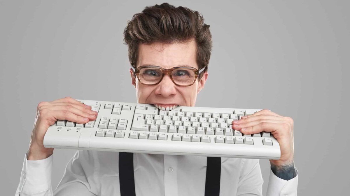 A geeky-looking young man with glasses bites down onto a computer keyboard in frustration or despair.