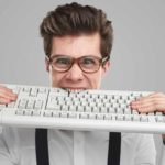 A geeky-looking young man with glasses bites down onto a computer keyboard in frustration or despair.