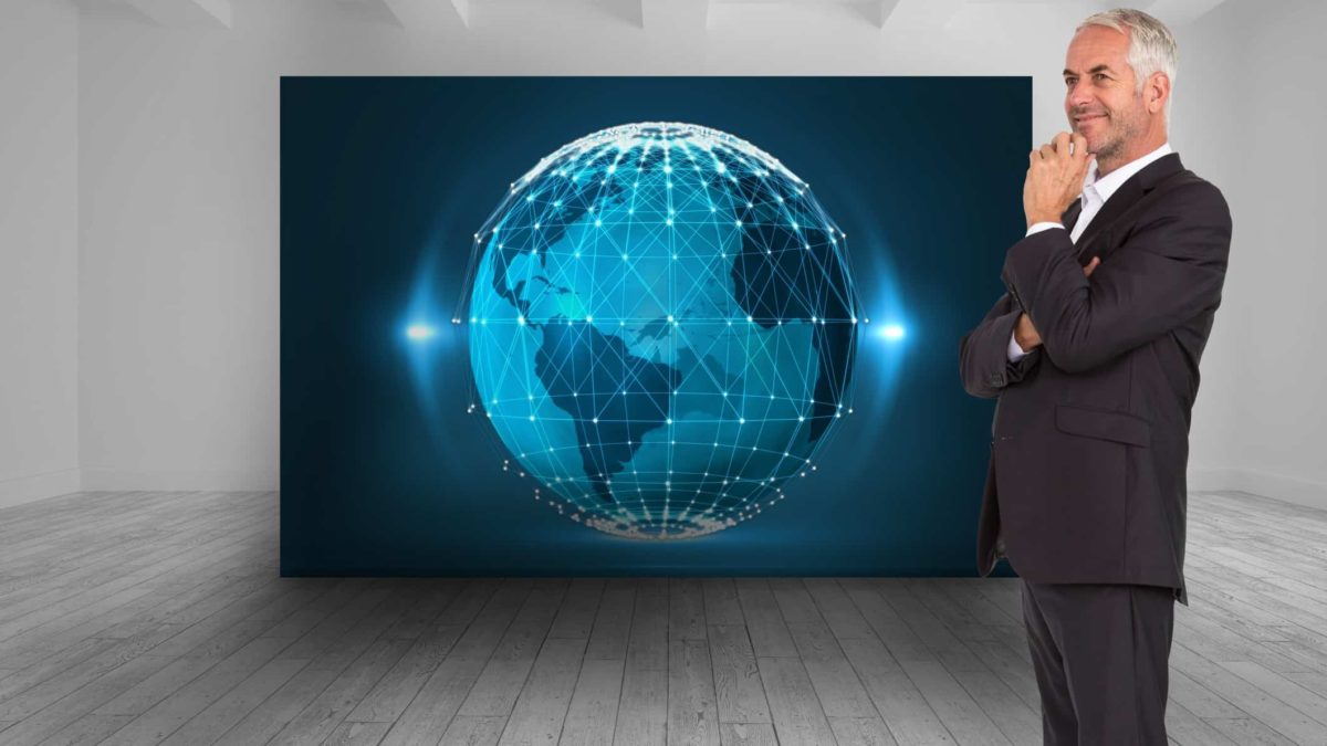 A man in a suit stands before a large backdrop of a blue-lit globe as the man smiles and holds his hand to his chin as though thinking.