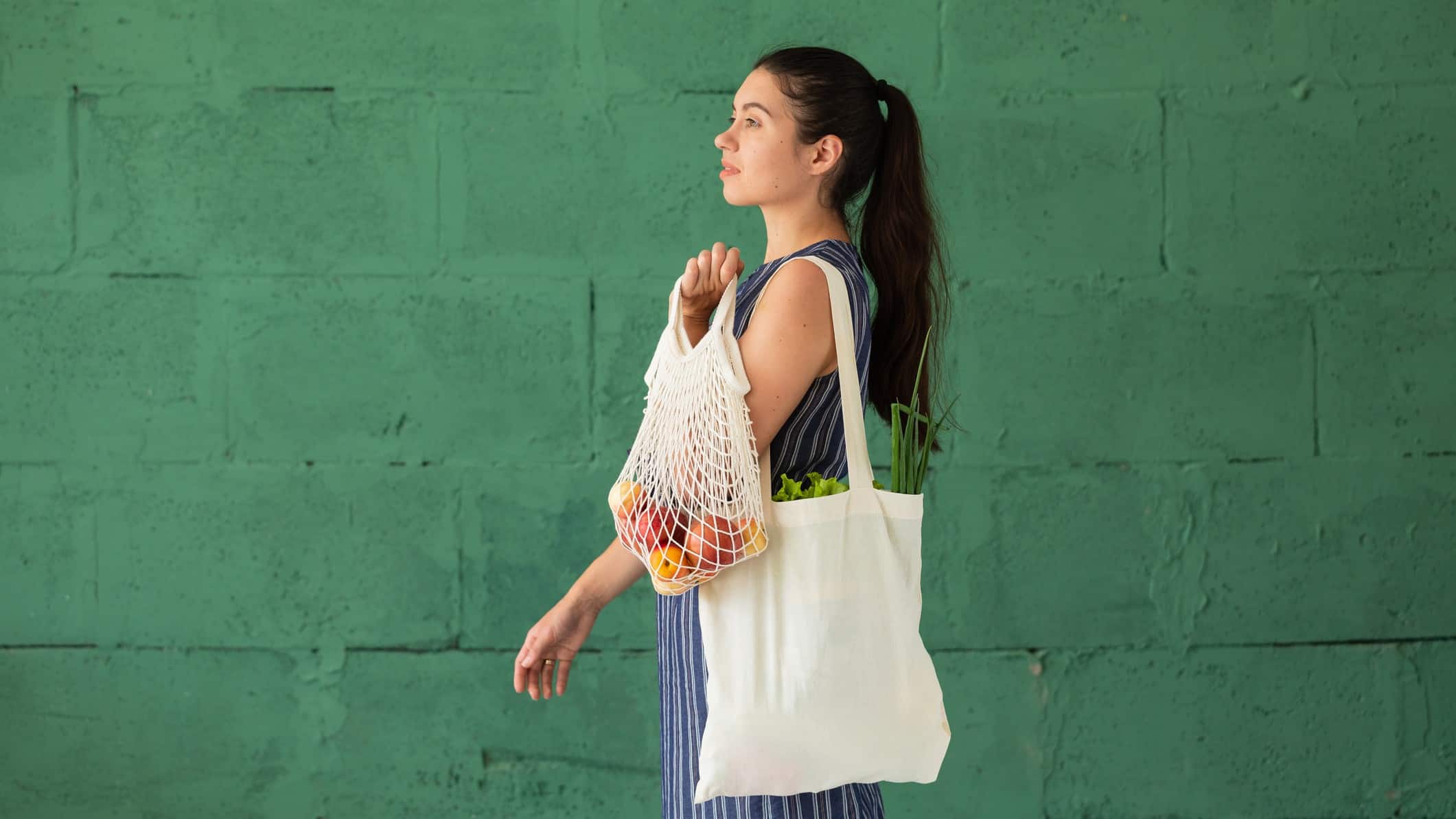 A woman carrying produce in recyclable shopping bags walks past a green wall, indicating consumer preference for sustainableretail