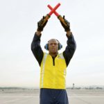 An airport ground staff worker holds two red beacons in either hand crossed above his head on a vast airport tarmac.