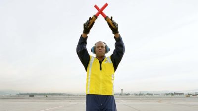An airport ground staff worker holds two red beacons in either hand crossed above his head on a vast airport tarmac.