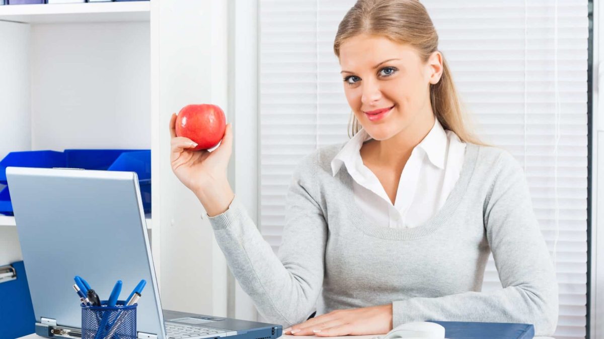 A woman in business attire sits at a desk in an office situation holding a red apple in her hand and smiling.