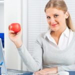 A smiling woman holds up an apple with a laptop open on her desk.