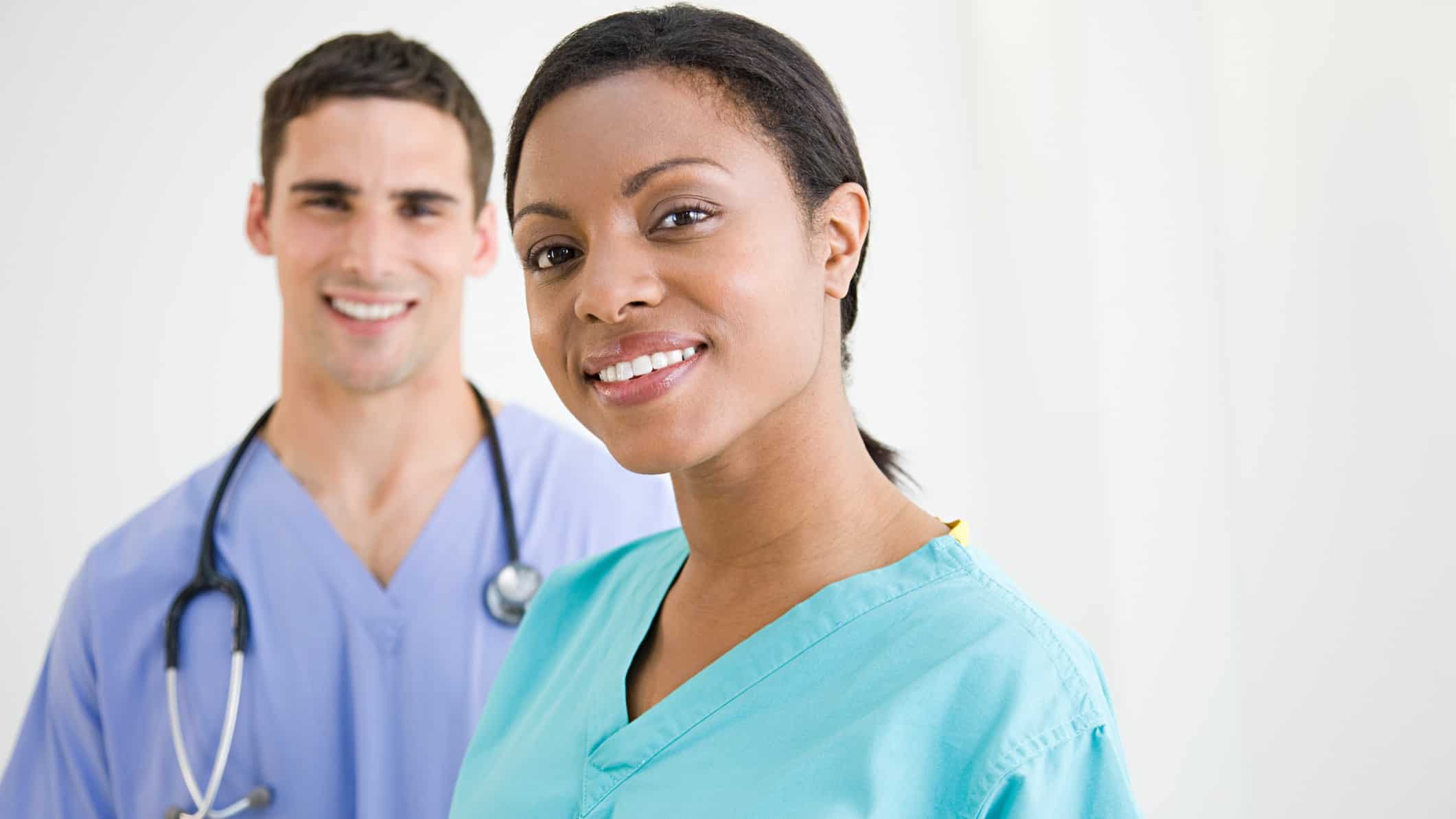 Two healthcare workers, a male doctor in the background with a woman in scrubs in the foreground,, smile towards the camera against a plain backdrop.