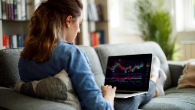 A casually dressed woman at home on her couch looks at index fund charts on her laptop
