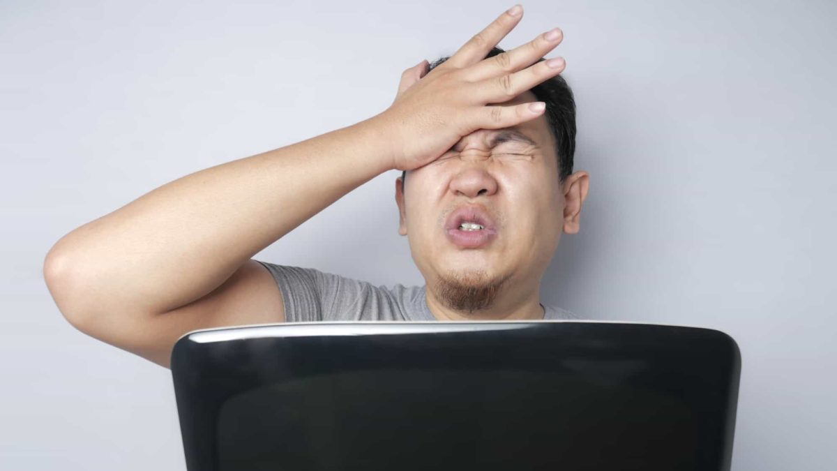 A young man clasps his hand to his head with his eyes closed and a pained expression on his face as he clasps a laptop computer in front of him, seemingly learning of bad news or a poor investment.