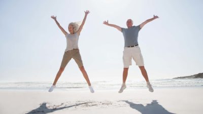 Two mature-age people, a man and a woman, jump in unison with their arms and legs outstretched on a sunny beach.