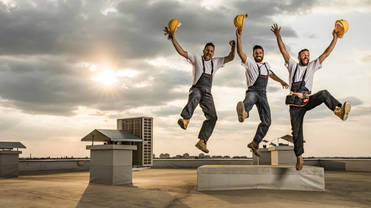 A group of three builders wearing worker overalls and carrying hard hats in their hands jumps jubilantly atopa rooftop space on a commercial building with an airconditioner shaft in the background and the sun behind a light cloud behind them.
