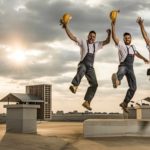 A group of three builders wearing worker overalls and carrying hard hats in their hands jumps jubilantly atopa rooftop space on a commercial building with an airconditioner shaft in the background and the sun behind a light cloud behind them.