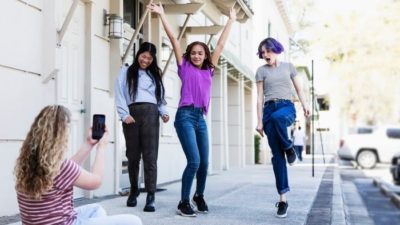 A group of four girls gather on a footapth in an urban setting with three of them in exuberant poses while the fourth girl uses her phone to take pictures of them.