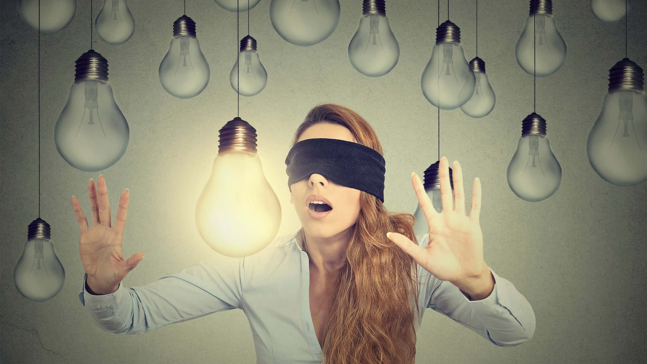 A young woman blindfolded holds her hands up as if feeling her way with the graphic of a lit up light bulb ahead of her and an assortment of unlit light bulbs hanging above her head.