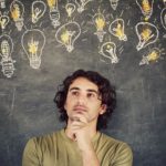 A young man looks like he his thinking holding his hand to his chin and gazing off to the side amid a backdrop of hand drawn lightbulbs that are lit up on a chalkboard.