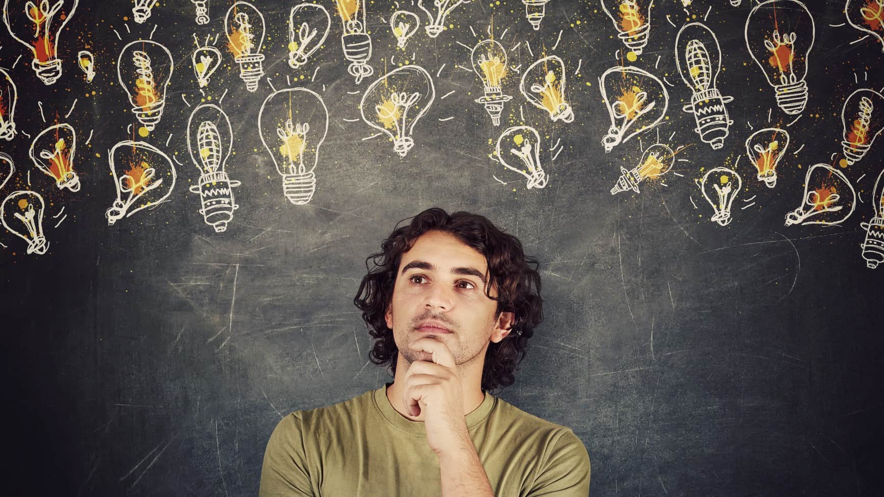 A young man looks like he his thinking holding his hand to his chin and gazing off to the side amid a backdrop of hand drawn lightbulbs that are lit up on a chalkboard.
