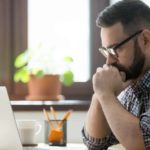 A man leans forward propped on his elbows as he holds his clasped hands to his mouth in a worried pose as he gazes at his computer screen in a home setting.