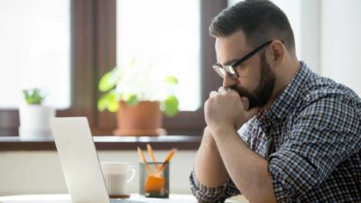 A man leans forward propped on his elbows as he holds his clasped hands to his mouth in a worried pose as he gazes at his computer screen in a home setting.