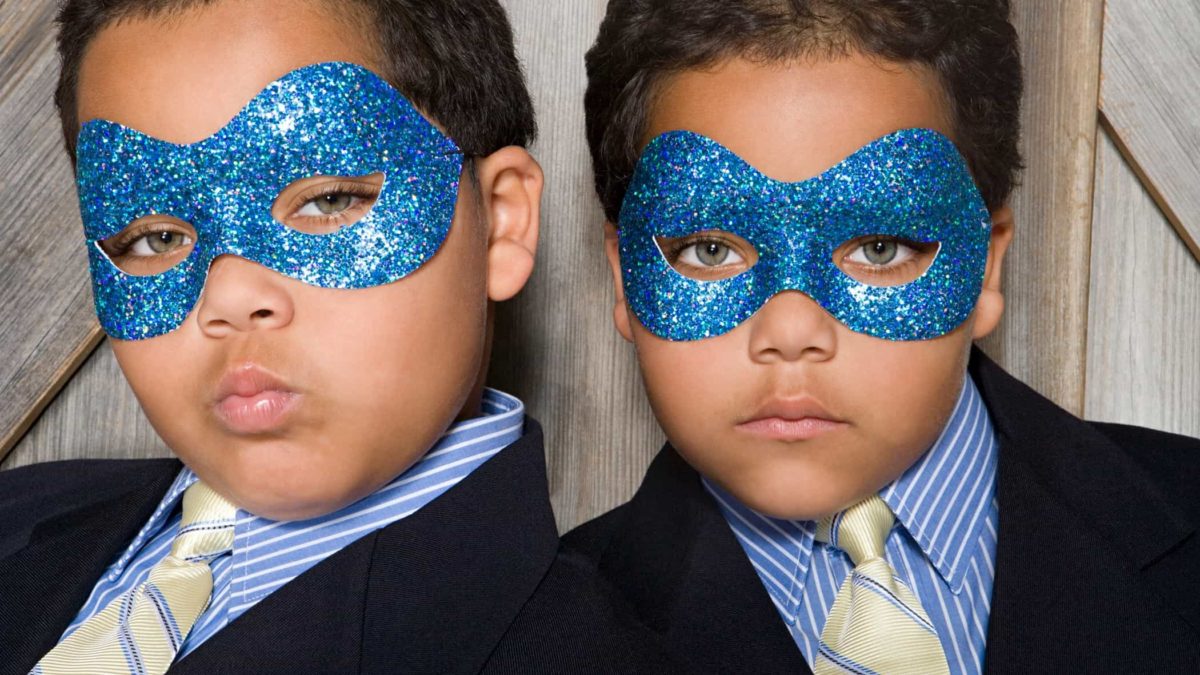 Two young boys, identical twins, dressed in suave business suits and ties wear sparkly masks over their eyes and pout at the camera.